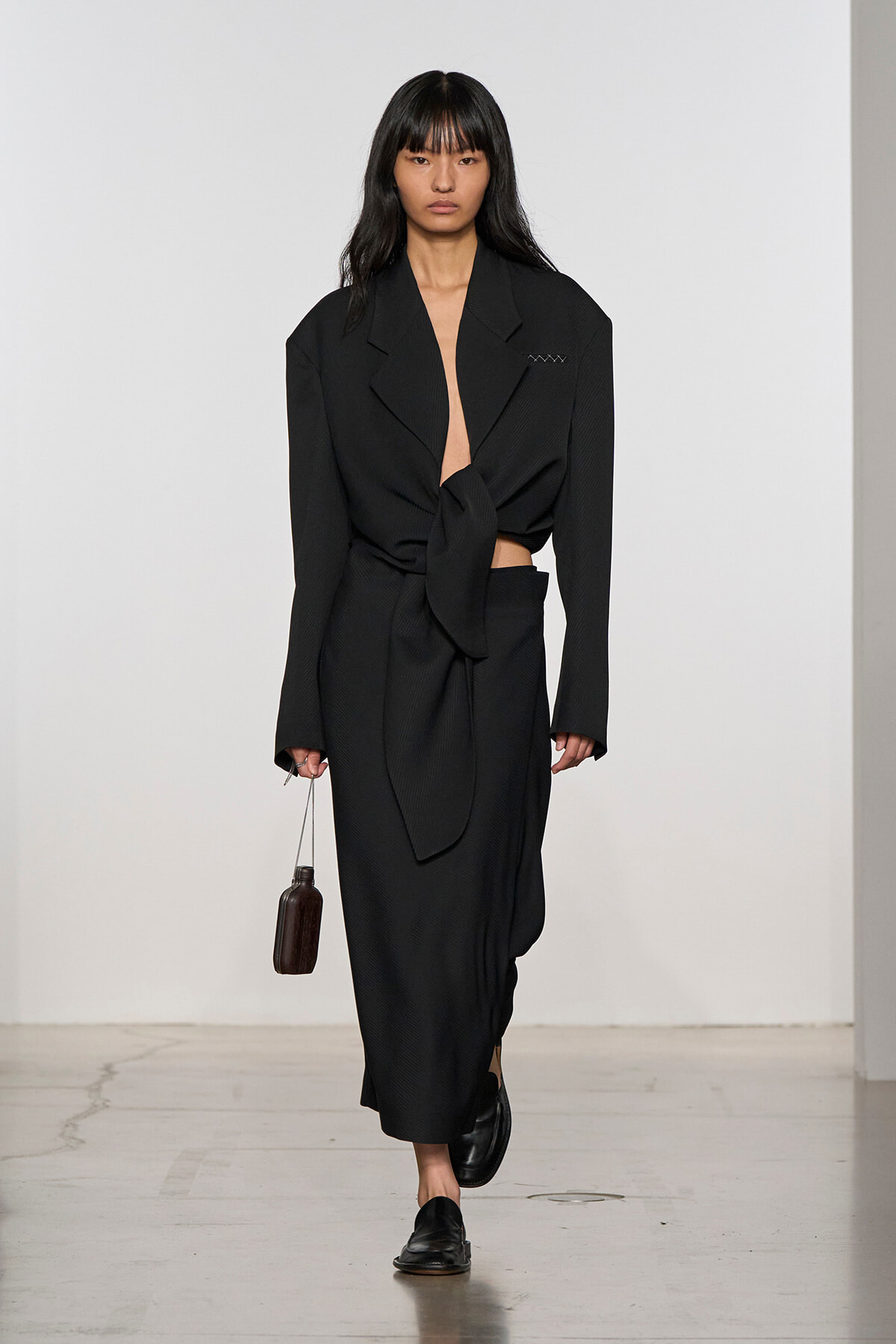 Model walking a runway in a black oversized blazer tied at the waist with a matching long skirt, carrying a small brown handbag.