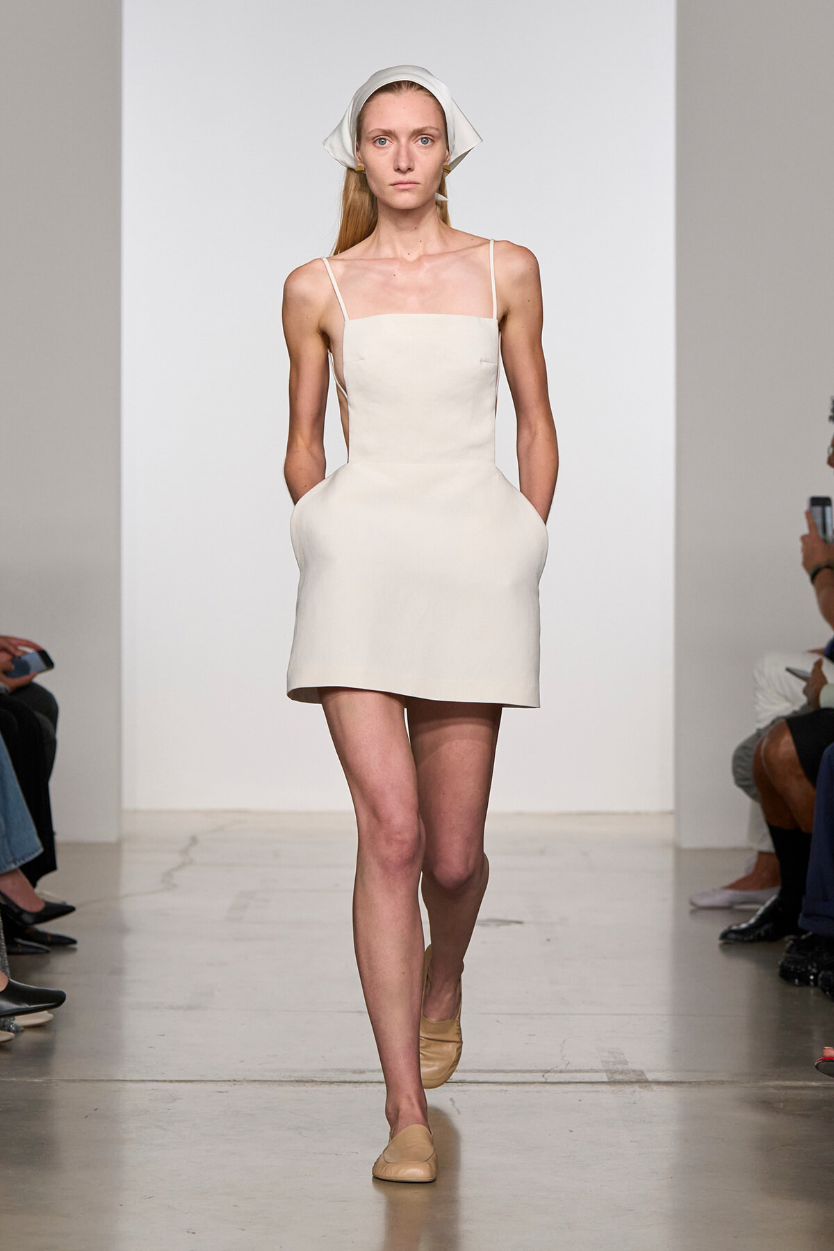 Model walking the runway in a minimalist ivory mini dress with thin straps and a white headscarf.