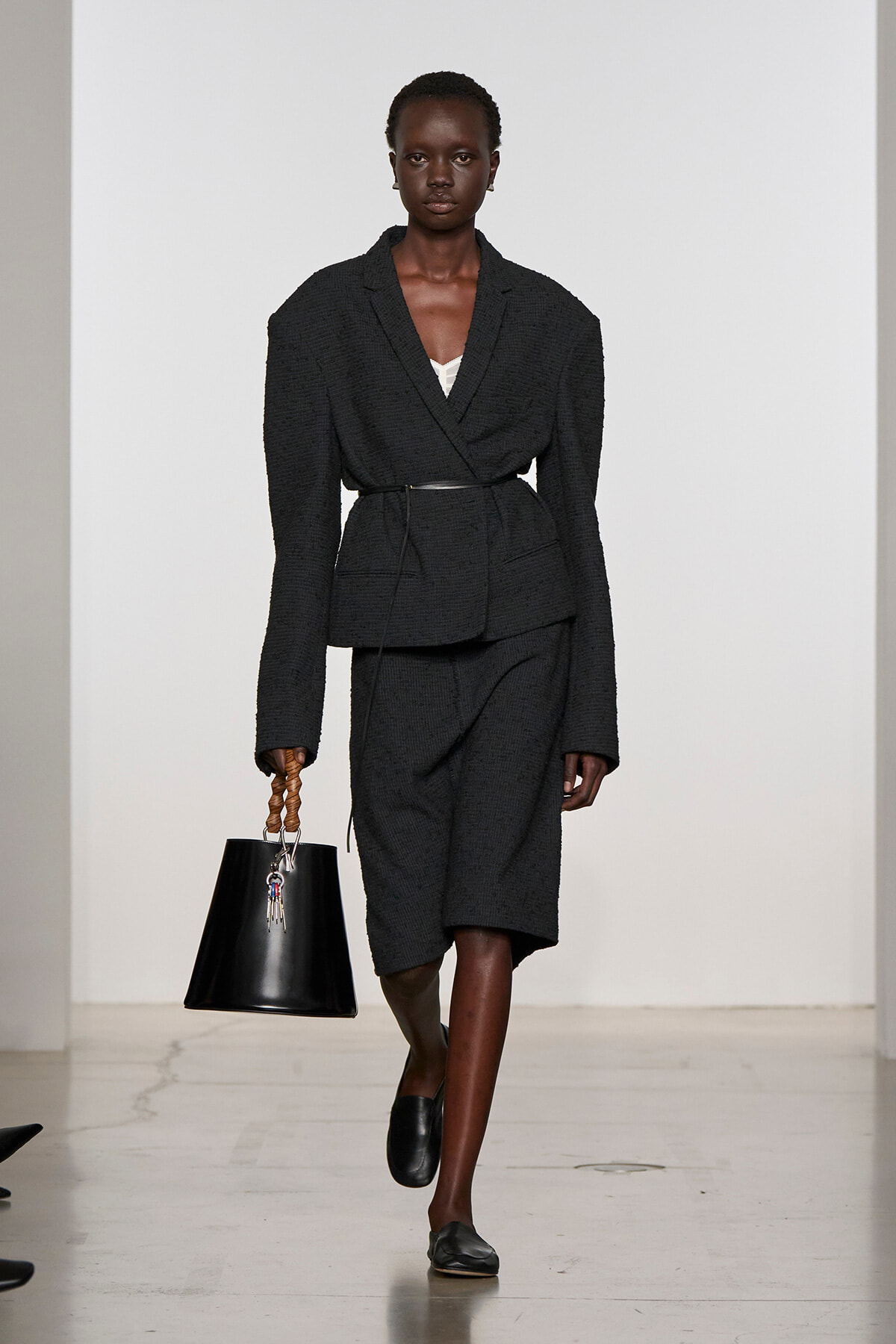 Model walking a fashion runway in a dark textured blazer and matching knee-length skirt with a belt, carrying a black handbag with a braided wooden handle.