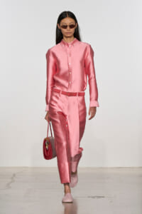 Model in pink satin button-down shirt and matching pants with a belt, carrying a red handbag and wearing narrow sunglasses on a runway.