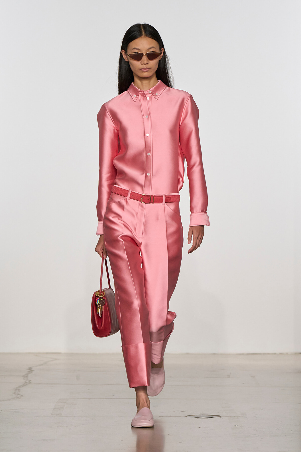Model in pink satin button-down shirt and matching pants with a belt, carrying a red handbag and wearing narrow sunglasses on a runway.
