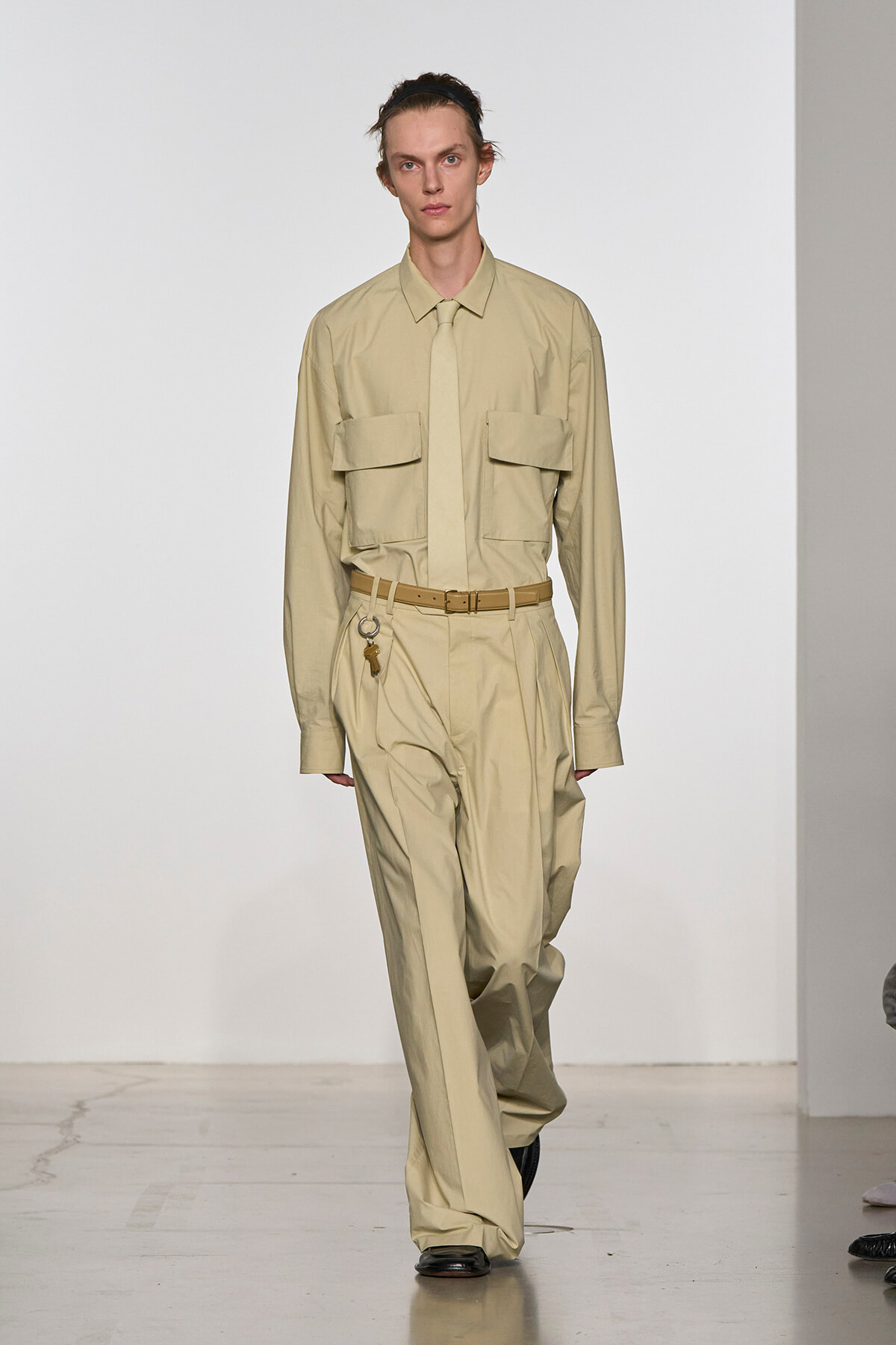 Male model on a runway wearing a beige utility jumpsuit with large front pockets and a matching belt.
