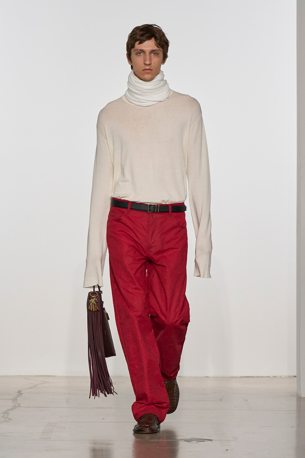 Male model walking a runway in a cream turtleneck sweater, red trousers, brown belt, and fringe handbag.