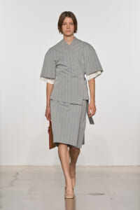Model walks the runway in a gray pinstriped kimono-style top with wide sleeves and a matching knee-length skirt, holding a brown clutch.