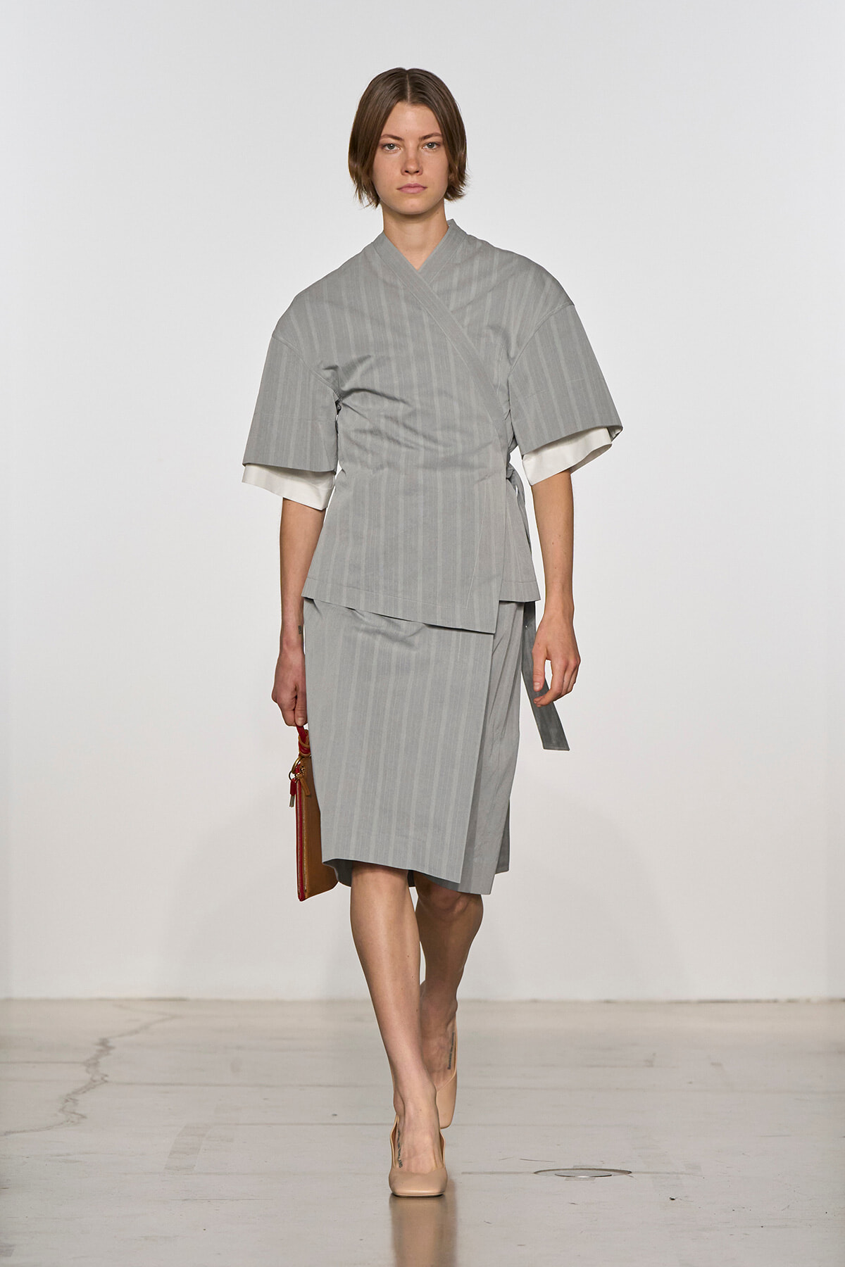 Model walks the runway in a gray pinstriped kimono-style top with wide sleeves and a matching knee-length skirt, holding a brown clutch.