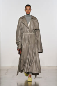 Model walking in a long taupe trench coat belted at the waist over a gray turtleneck, paired with yellow sneakers and a small burgundy handbag.