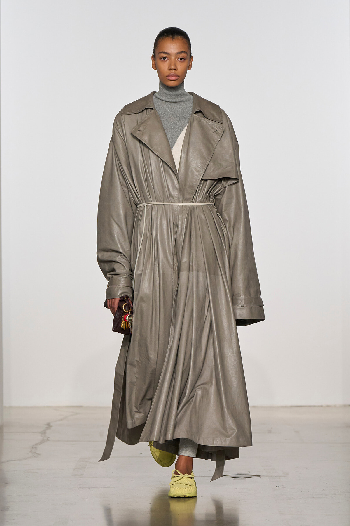 Model walking in a long taupe trench coat belted at the waist over a gray turtleneck, paired with yellow sneakers and a small burgundy handbag.