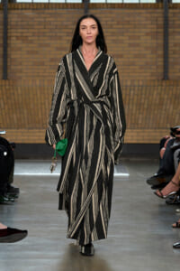 Model walks the runway in a black-and-cream striped belted robe-like coat, holding a small green clutch with keys attached