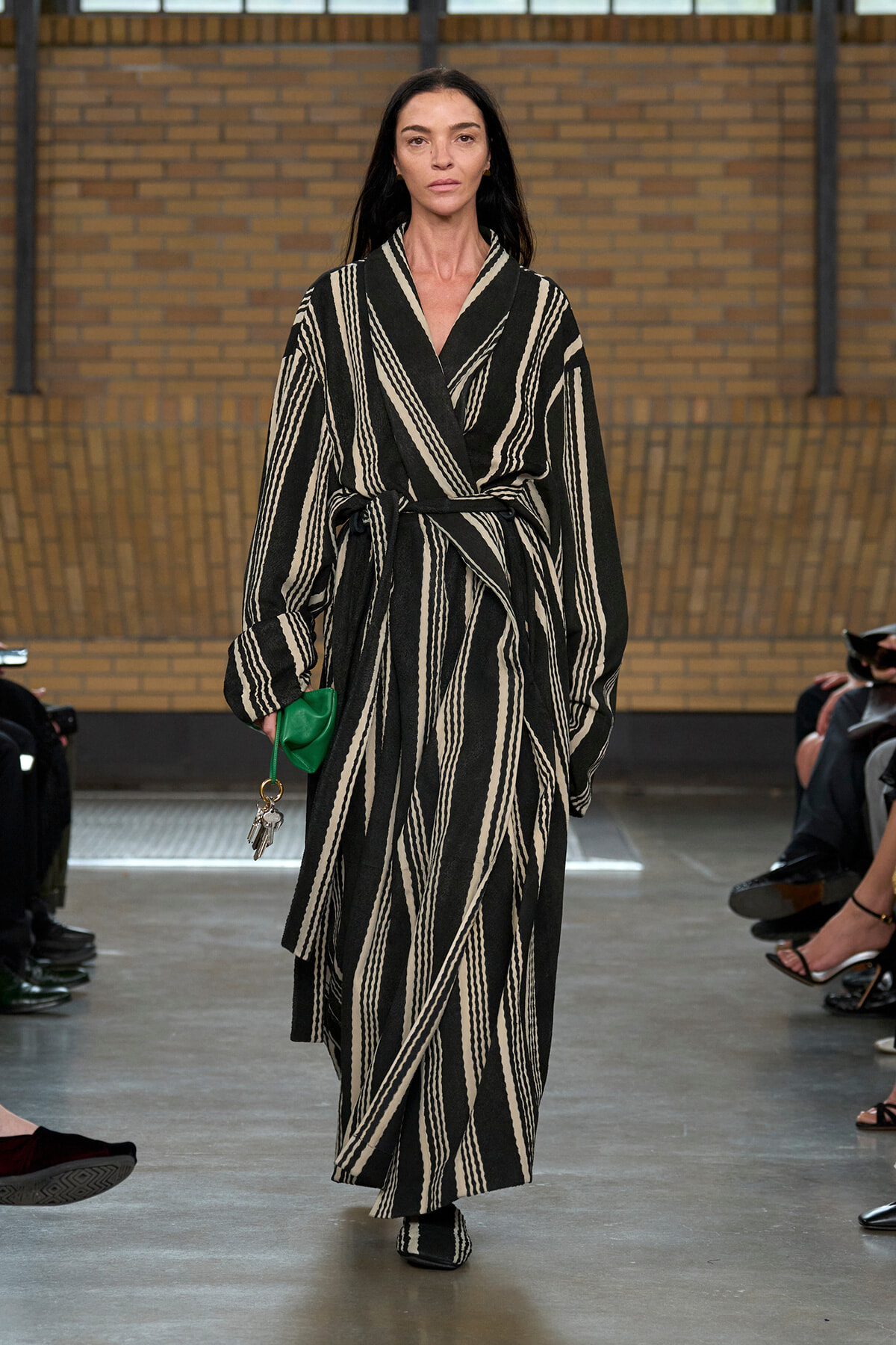 Model walks the runway in a black-and-cream striped belted robe-like coat, holding a small green clutch with keys attached