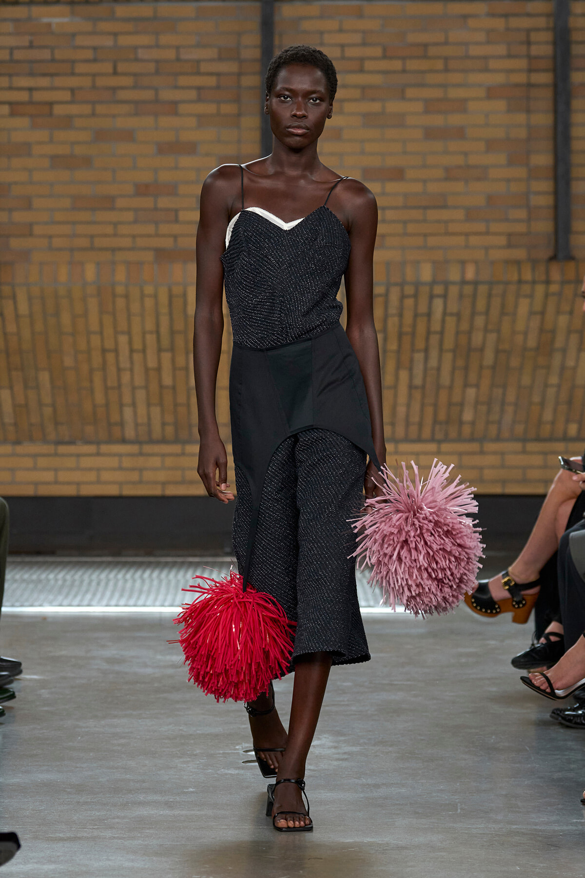 Model on a runway wearing a black glittery top with an asymmetrical draped bottom, carrying oversized red and pink fluffy pom-pom bags.