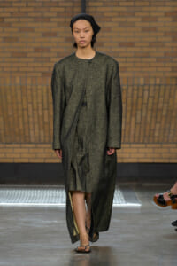 Model walking a runway in a long, dark grey textured coat with a black headscarf.