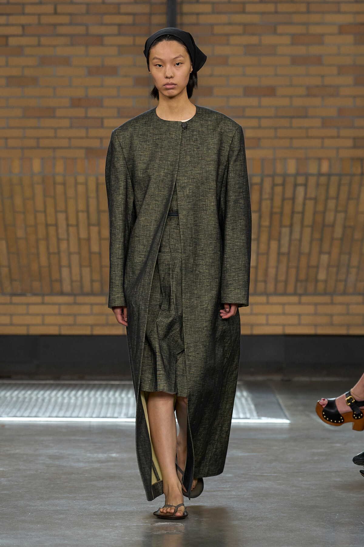 Model walking a runway in a long, dark grey textured coat with a black headscarf.