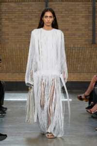 Model on a runway wearing a white fringe gown with long vertical fringe strips and a matching fringe bag by her side.
