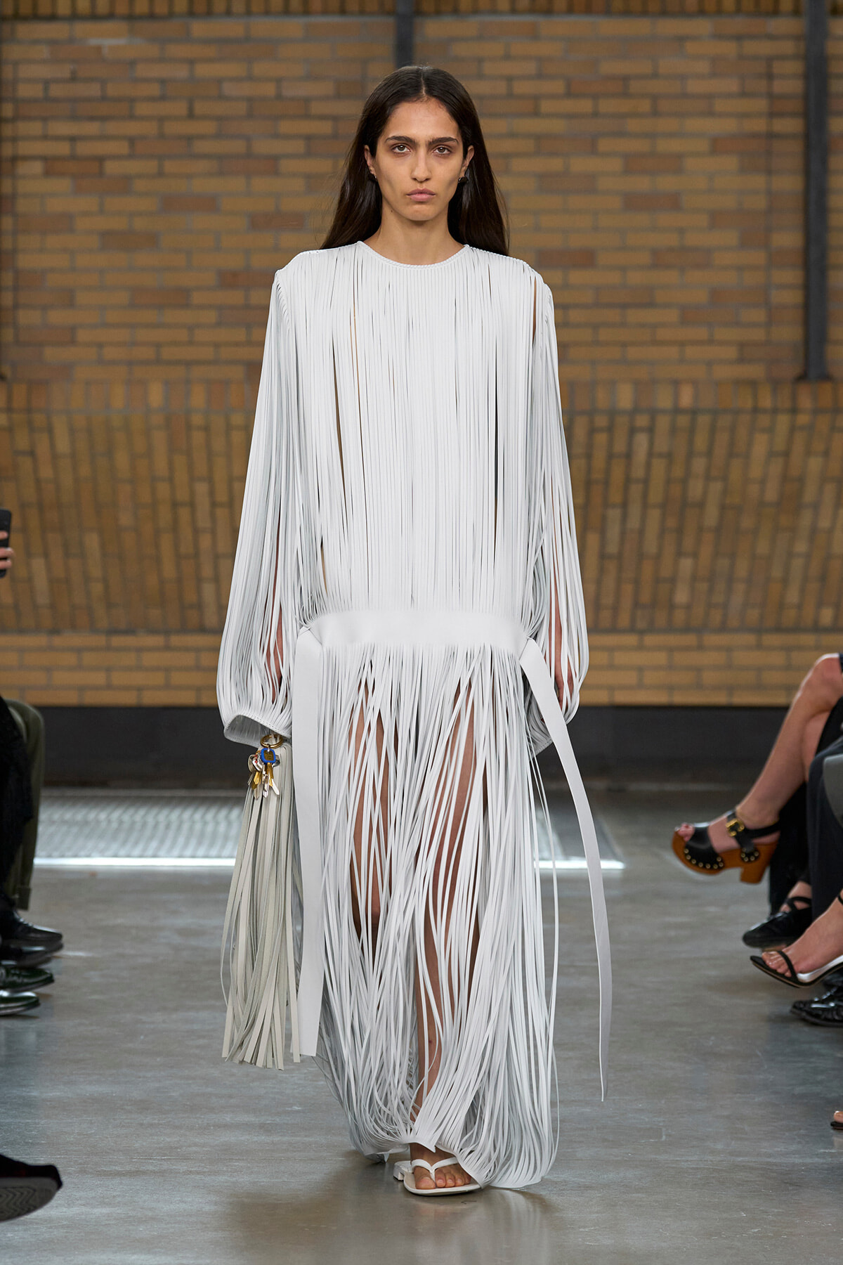 Model on a runway wearing a white fringe gown with long vertical fringe strips and a matching fringe bag by her side.