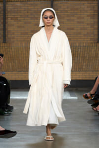 Model wearing a white bathrobe with a belt and hood, sunglasses, and white sandals walking a fashion show runway.