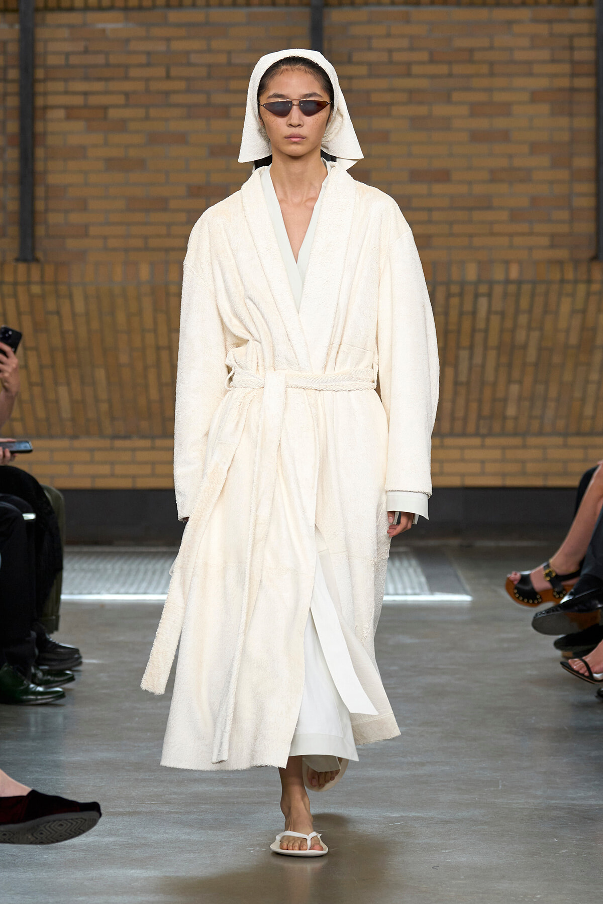 Model wearing a white bathrobe with a belt and hood, sunglasses, and white sandals walking a fashion show runway.