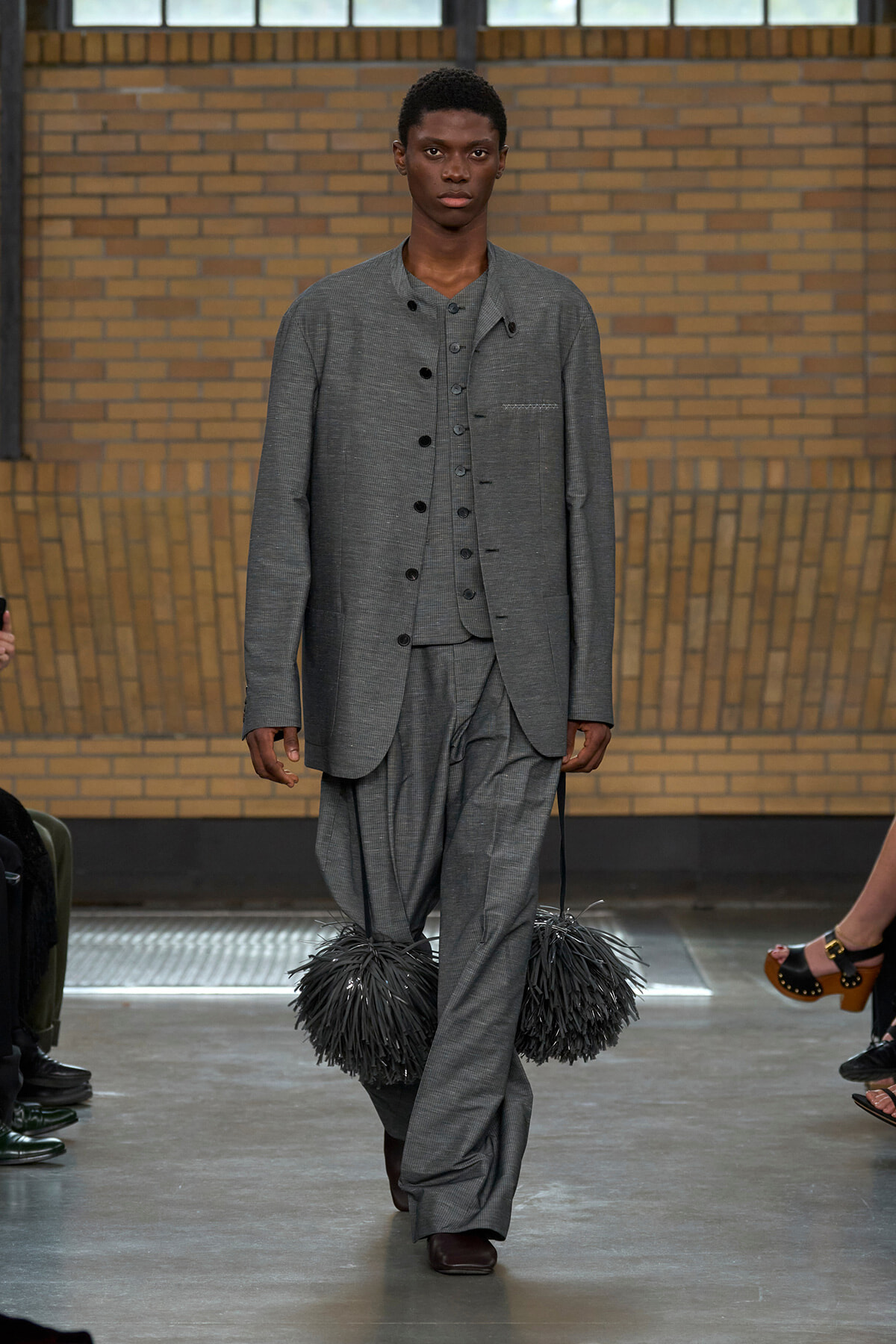 Male model walks a runway in a gray three-piece suit with oversized trousers, carrying two black fluffy pom-pom bags at the sides.