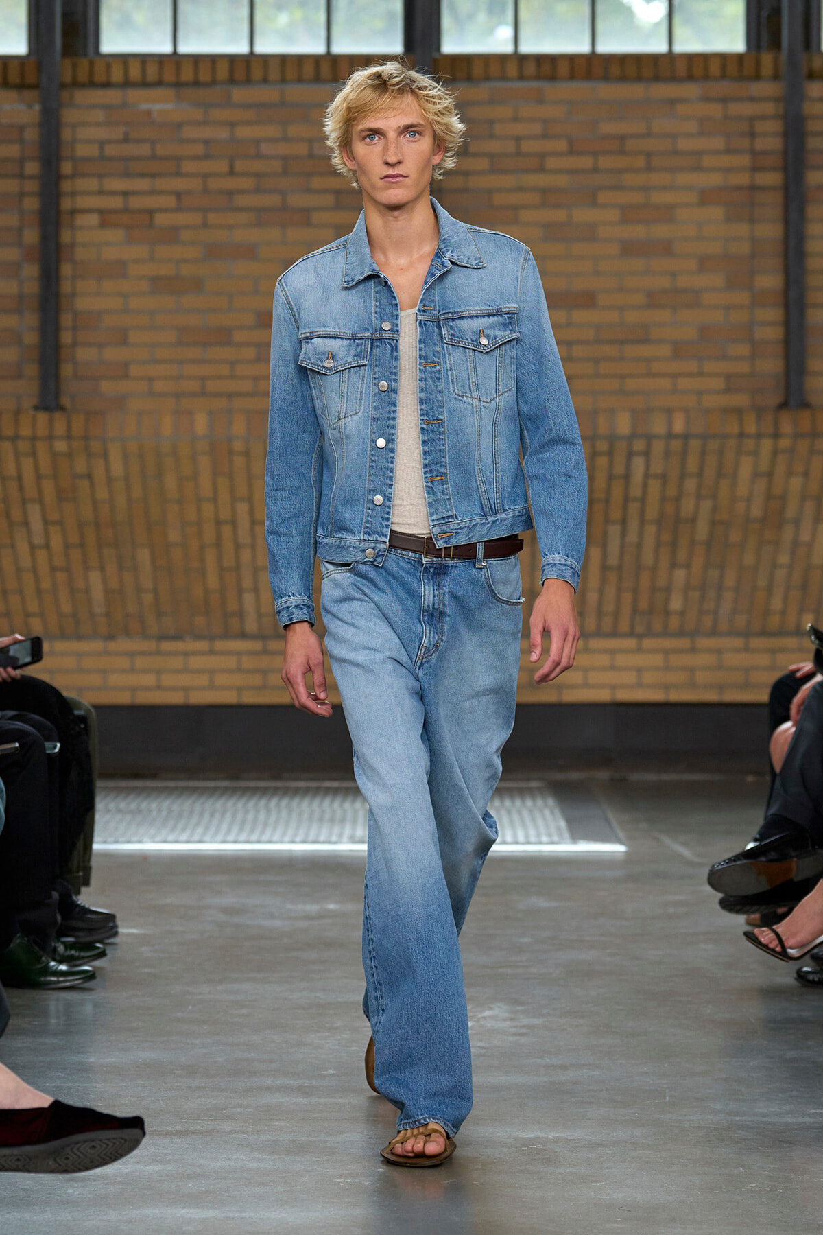 Male model walks the runway in a light denim jacket and wide-leg jeans with a tan belt and sandals, brick wall backdrop.