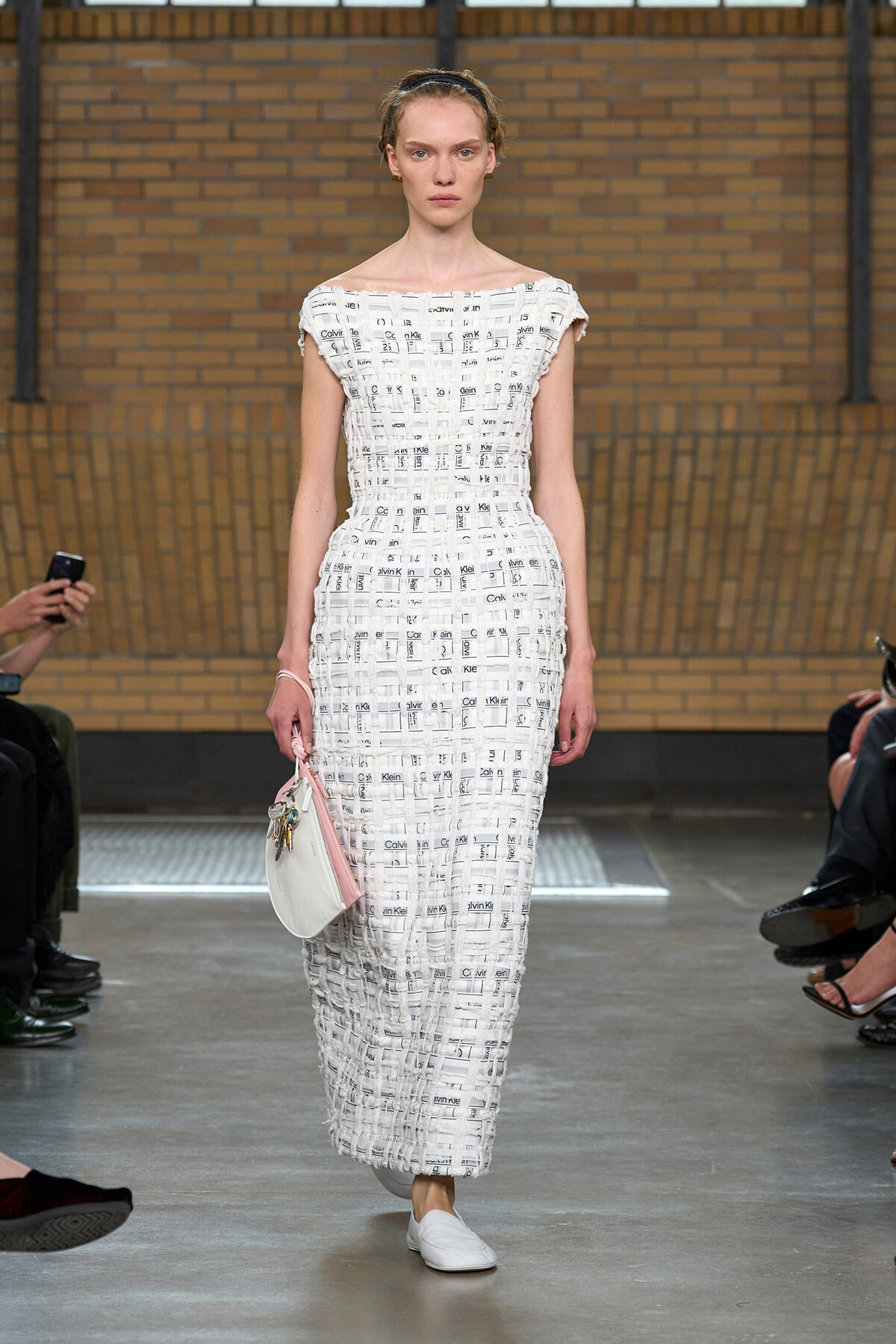 Full-length shot of a model walking a runway in a white, textured, off-the-shoulder maxi dress with a woven, print-like pattern, carrying a small white and pink handbag and wearing white loafers.