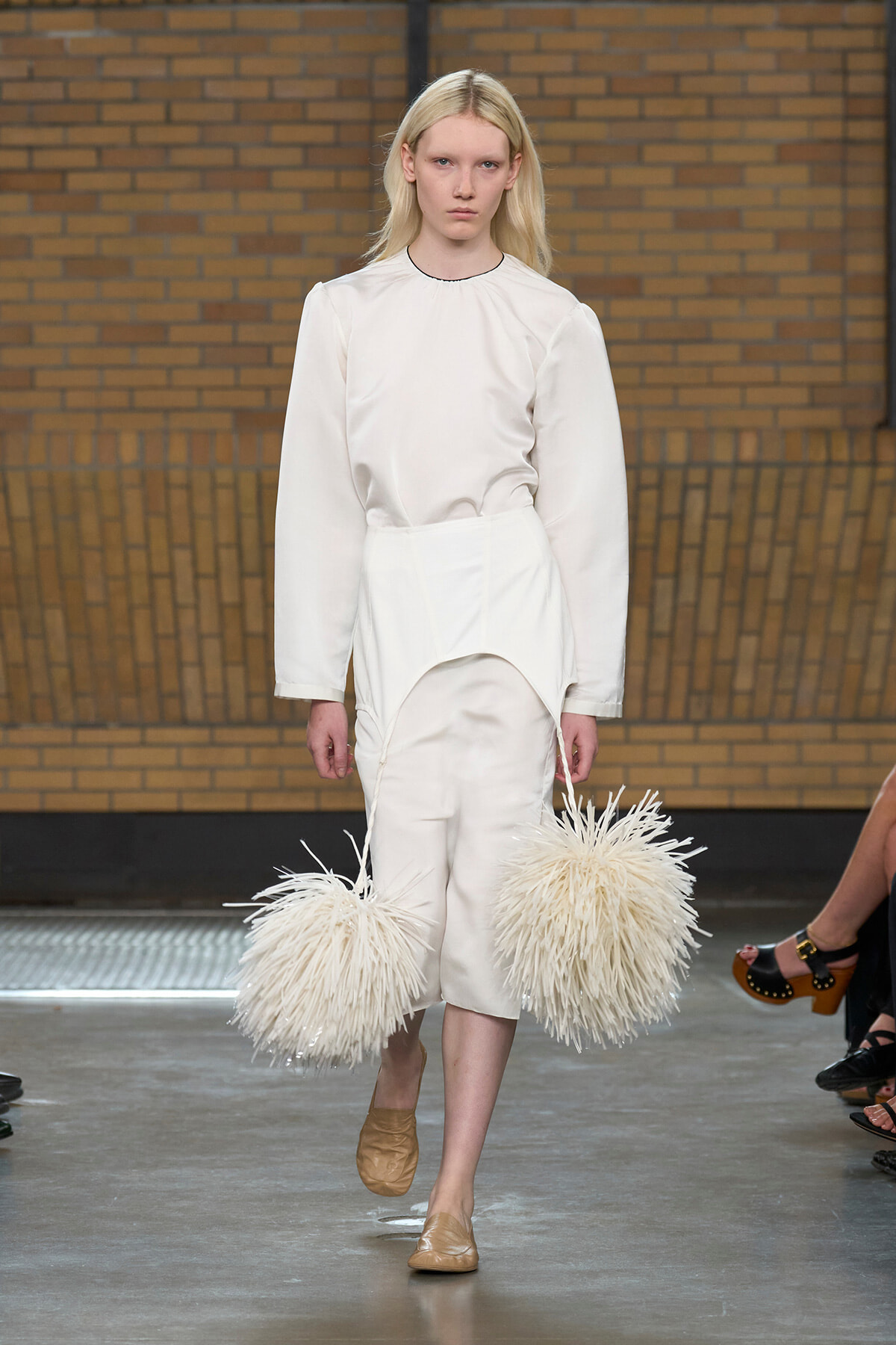 Model on a runway wearing an all-white outfit with two large cream shaggy pom-poms hanging from strings at her sides.