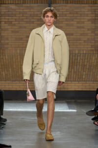 Male model wearing a light beige jacket over a cream striped shirt and matching striped shorts, carrying a small pink handbag, walking on a runway.