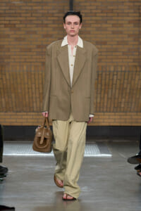 Model on a runway wearing an oversized tan blazer, white shirt, beige wide-leg pants, brown sandals, and carrying a brown handbag against a brick backdrop.
