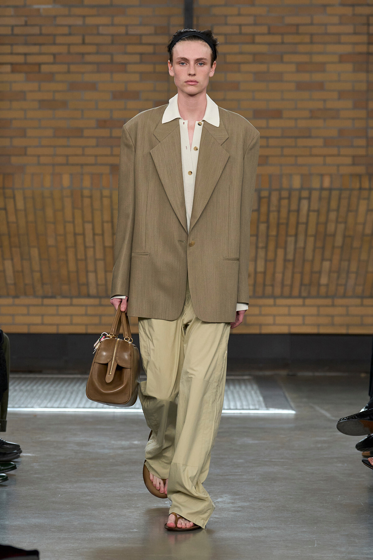 Model on a runway wearing an oversized tan blazer, white shirt, beige wide-leg pants, brown sandals, and carrying a brown handbag against a brick backdrop.