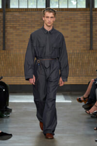 Man walking a fashion runway wearing a dark gray utility jumpsuit with a slim belt and brown shoes.