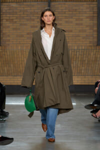 Model walking the runway in an oversized olive trench coat belted at the waist, with a white shirt and blue jeans, carrying a green handbag.