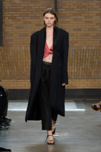 Model walks a runway in a long black overcoat over a red textured crop top and black wide-leg pants, wearing strappy sandals.