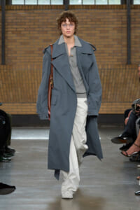 Model walking a runway in an oversized blue-gray trench coat over a gray shirt, white cargo pants, and a brown shoulder bag. She wears glasses and looks forward.