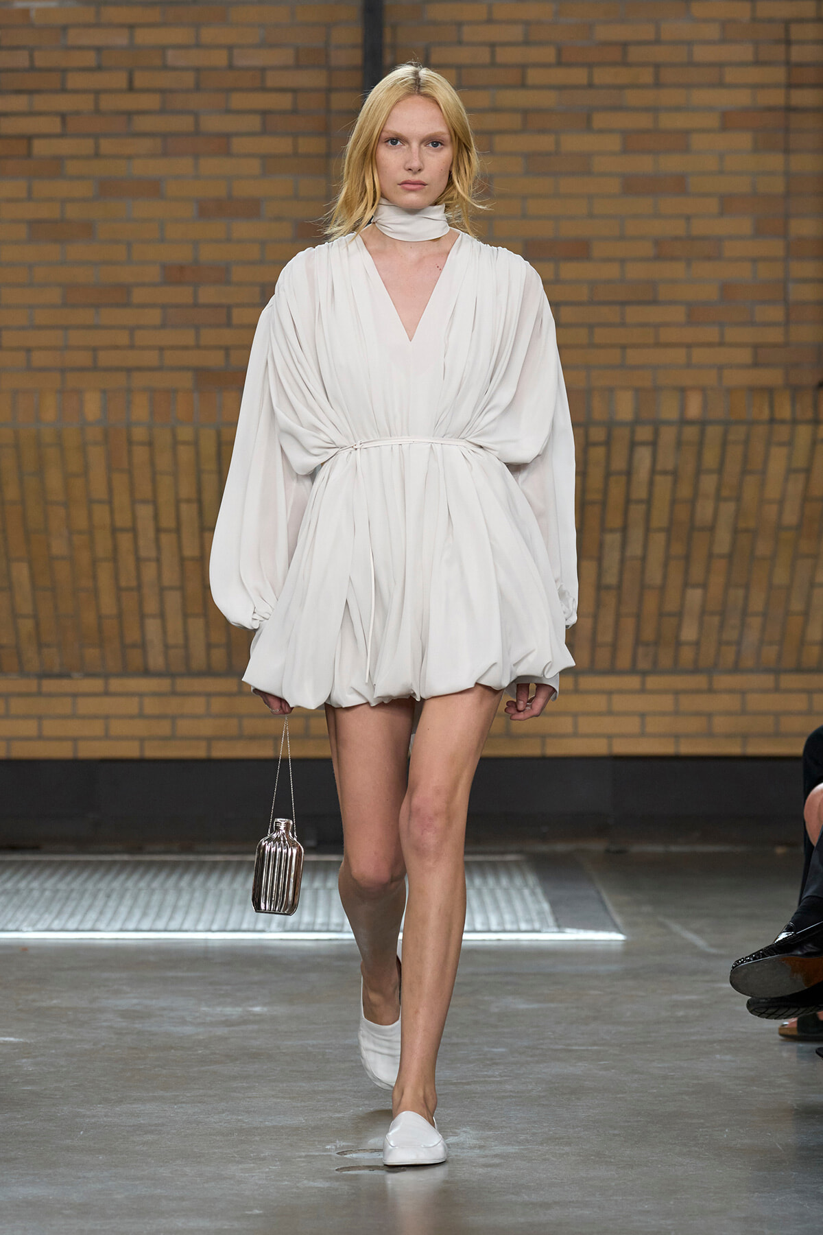 Model walking runway in a white draped V-neck dress with voluminous sleeves and a gathered bubble skirt, holding a small metallic handbag.