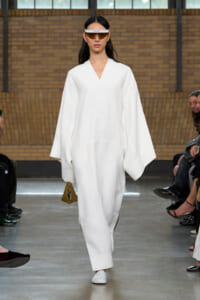Model on a runway wearing an oversized white robe-like outfit, white flats, a white visor, and carrying a small tan handbag with a blue charm.