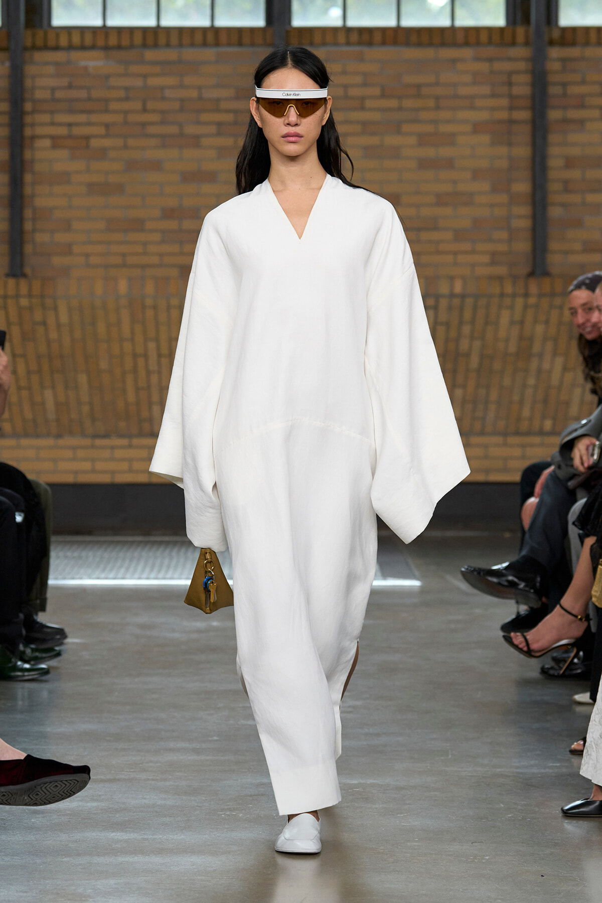 Model on a runway wearing an oversized white robe-like outfit, white flats, a white visor, and carrying a small tan handbag with a blue charm.