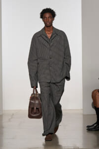 Male model walking the runway in a gray checkered suit, carrying a brown leather briefcase.