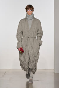Male model on a runway wearing a crinkled light-gray wrap coat belted at the waist, with a gray turtleneck and sunglasses, holding a small red pouch.