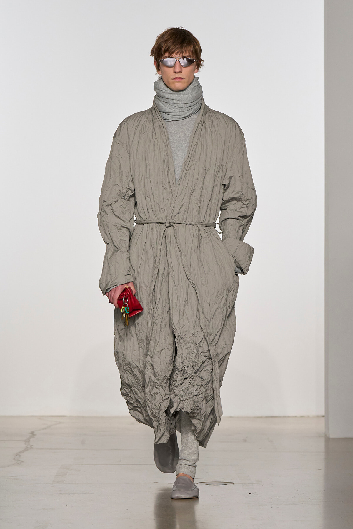 Male model on a runway wearing a crinkled light-gray wrap coat belted at the waist, with a gray turtleneck and sunglasses, holding a small red pouch.