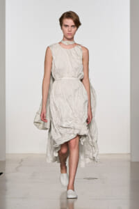 Model walking a fashion runway in a pale ivory, crinkled sleeveless dress with a belt and cape-like back piece.