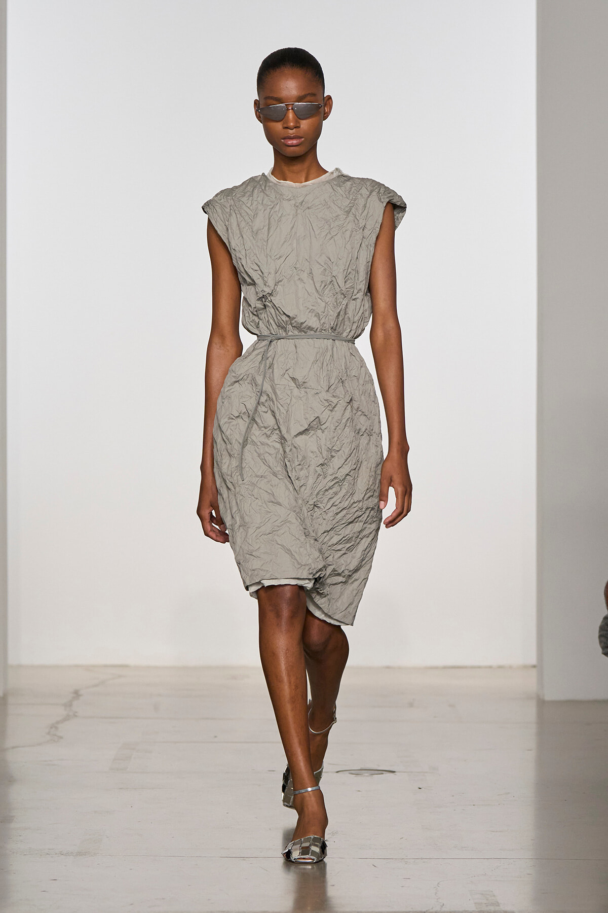 Female model walking a runway in a wrinkled light-gray belted dress with short sleeves and narrow sunglasses.