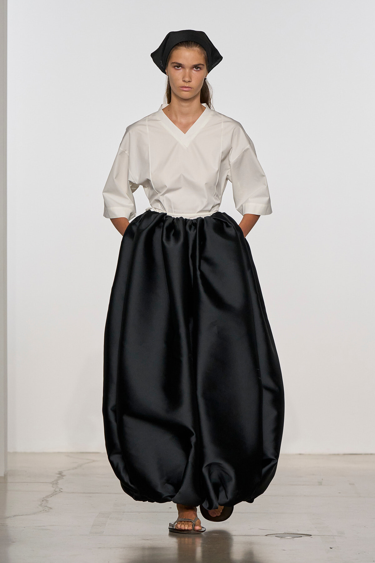 Model walking a runway in a white V-neck blouse paired with a voluminous black balloon skirt, black head wrap, and sandals.
