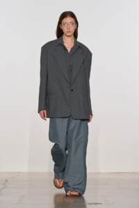 Model on a runway wearing an oversized dark gray blazer over a gray shirt and baggy blue-gray pants, sandals.