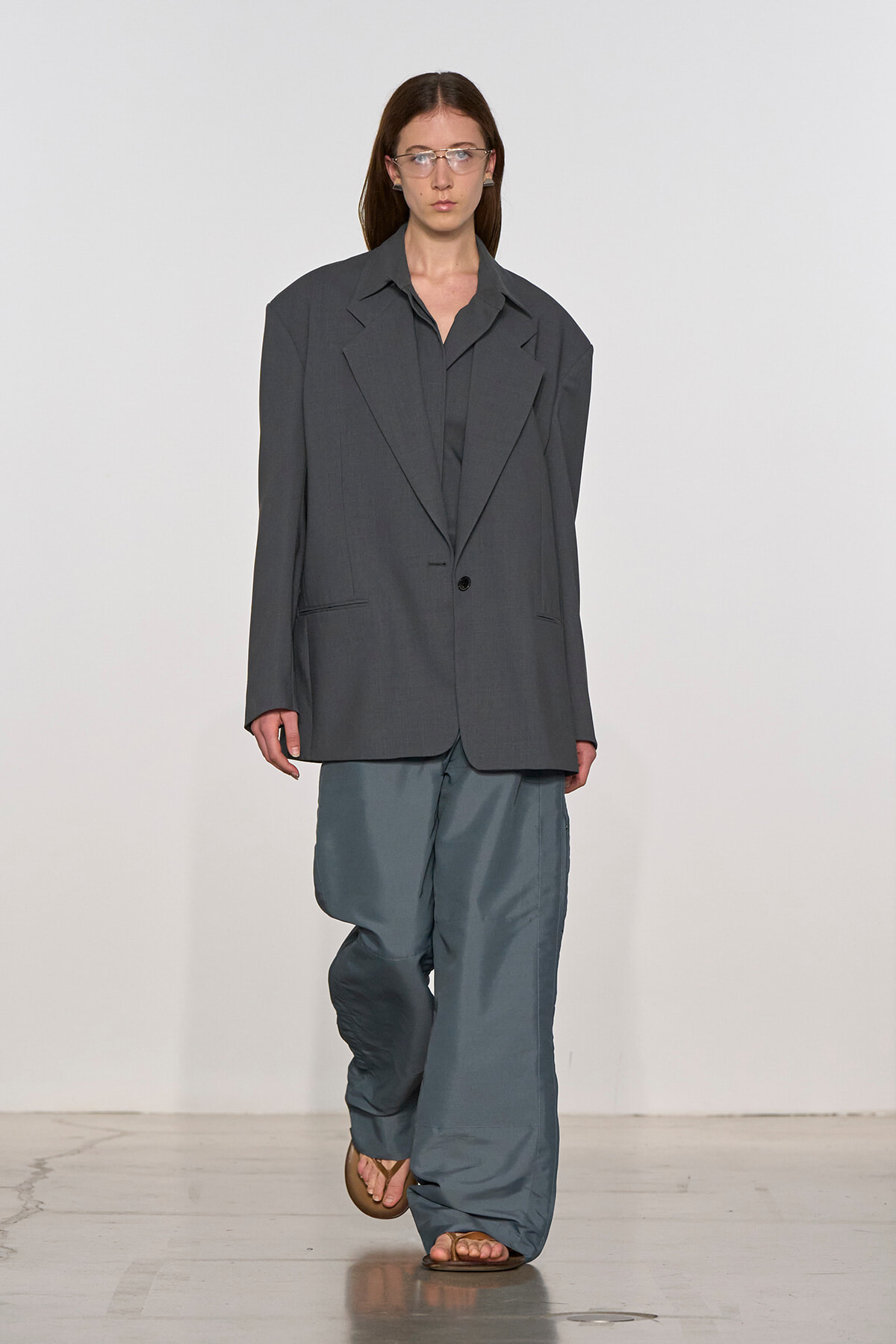 Model on a runway wearing an oversized dark gray blazer over a gray shirt and baggy blue-gray pants, sandals.