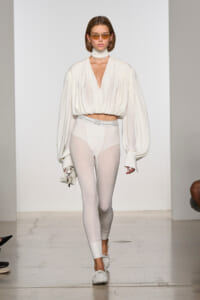 Runway model in an all-white look: draped blouse with a deep V, white leggings, and white sneakers, carrying a small white bag with keys.