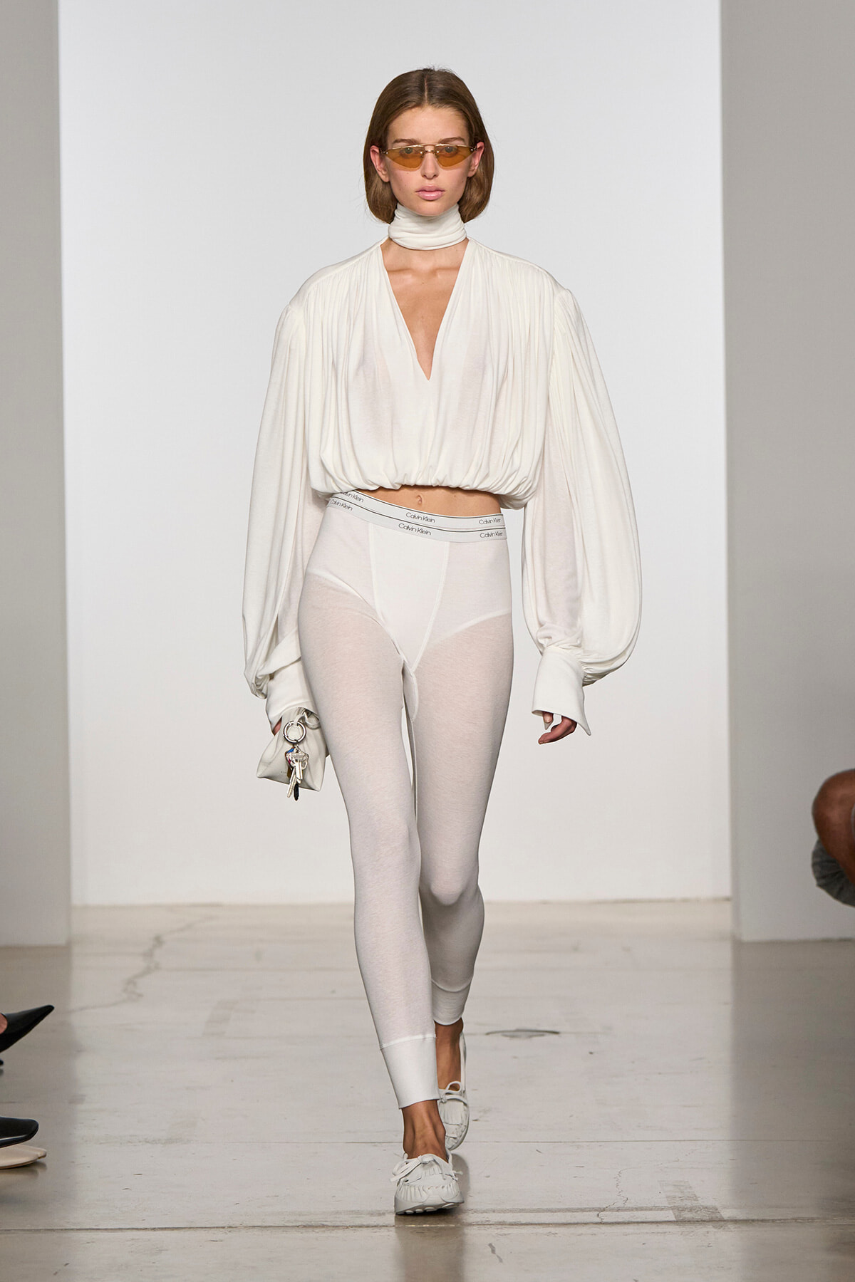 Runway model in an all-white look: draped blouse with a deep V, white leggings, and white sneakers, carrying a small white bag with keys.
