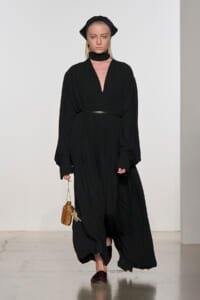 Runway model in a long black pleated dress with a thin belt, head wrap, and scarf, carrying a small wooden handbag and keys.