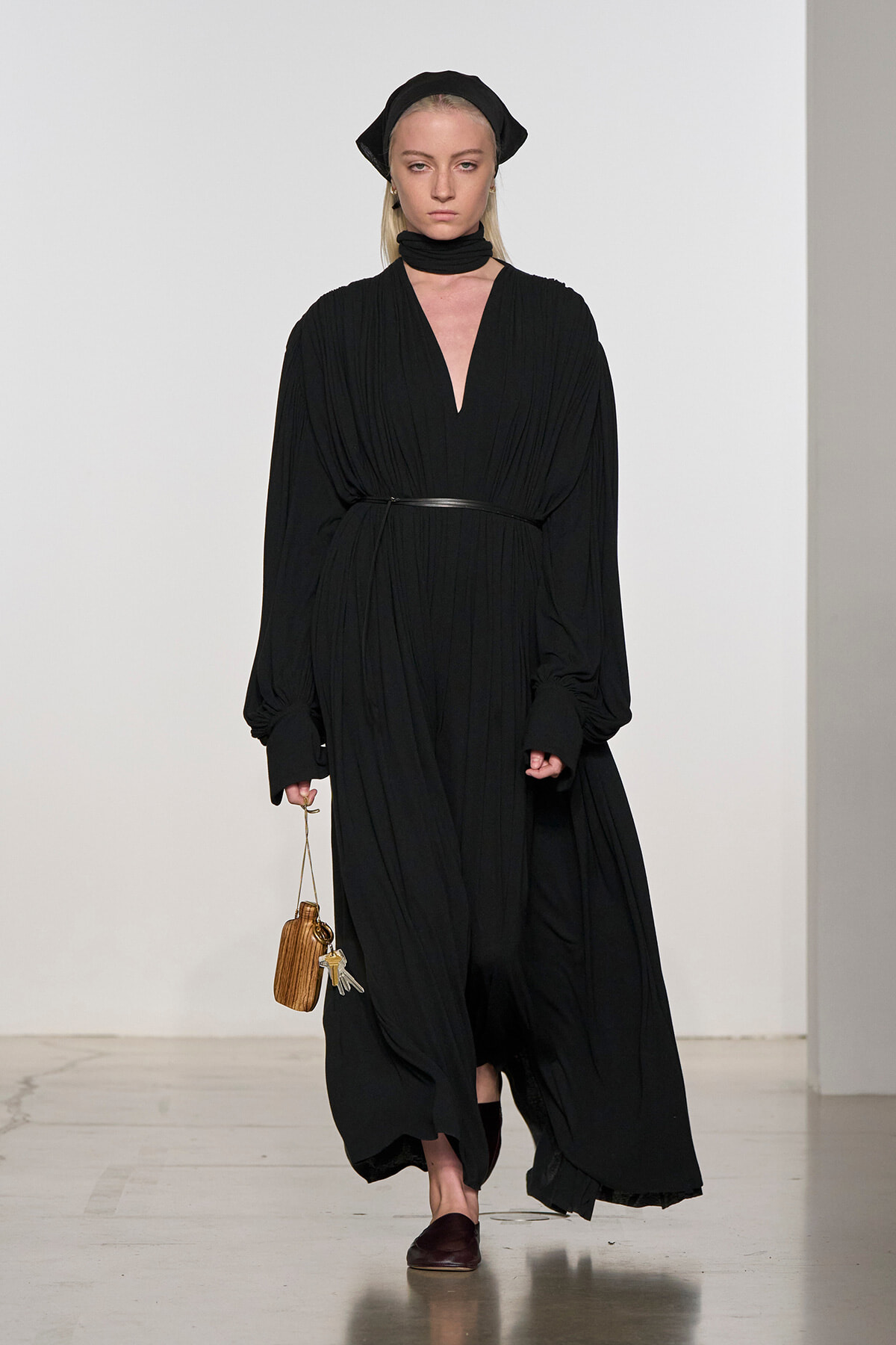 Runway model in a long black pleated dress with a thin belt, head wrap, and scarf, carrying a small wooden handbag and keys.