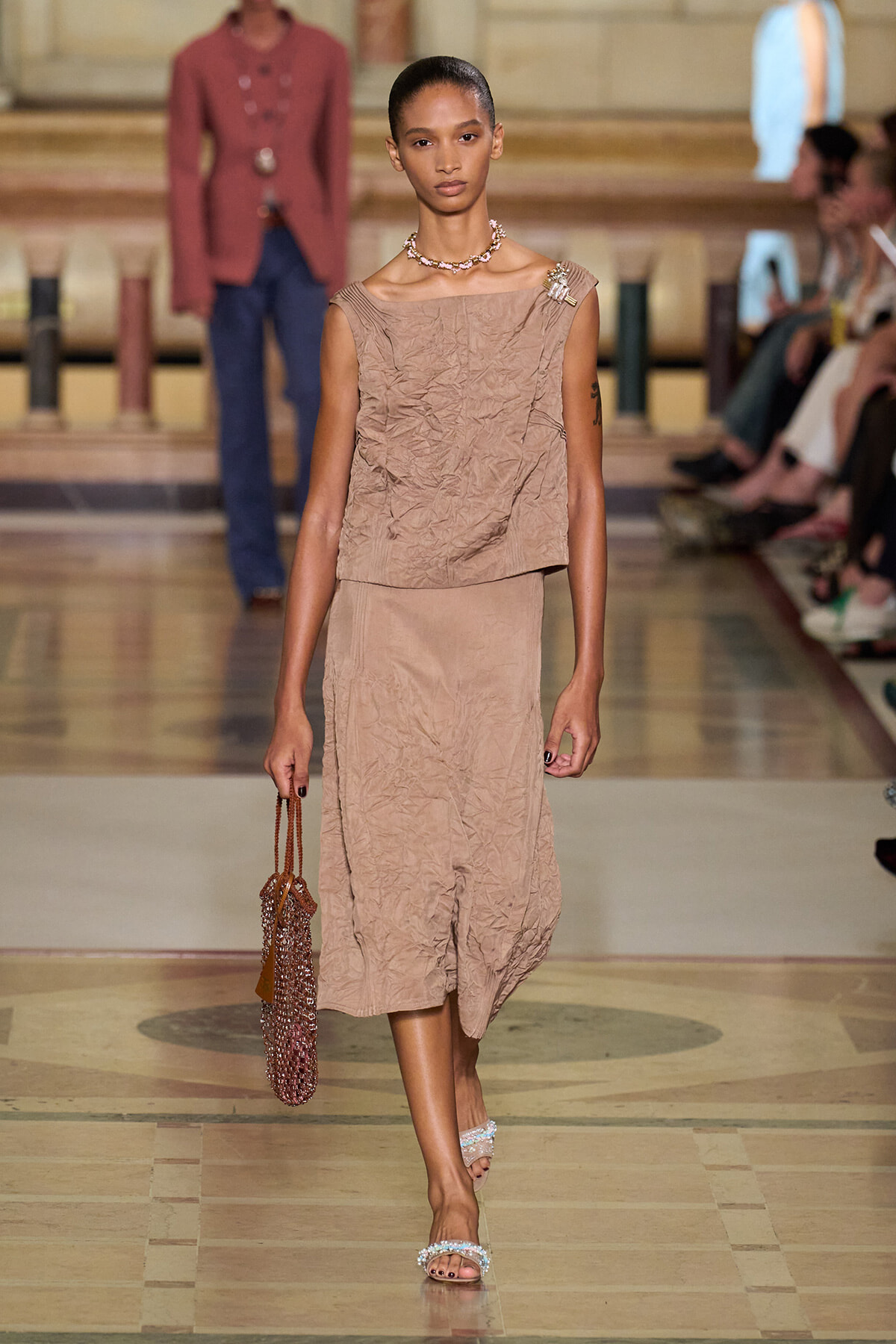 Model walks the runway in a crinkled beige sleeveless top and matching midi skirt, accessorized with a necklace and a beaded handbag.
