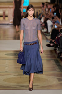 Model walks a runway in a gray knit top tucked into a blue midi skirt, accessorized with a brown belt and blue handbag as she strides forward.