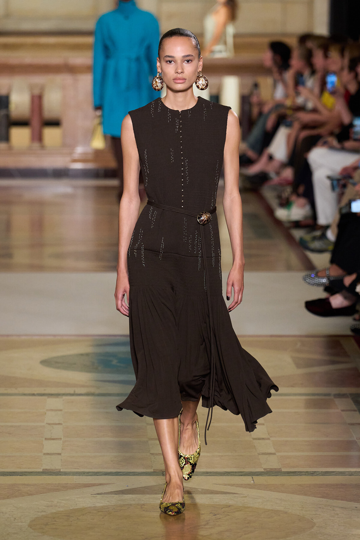 Female model walking a runway in a sleeveless brown dress with beaded details and a waist belt, paired with large circular earrings and snakeskin heels.