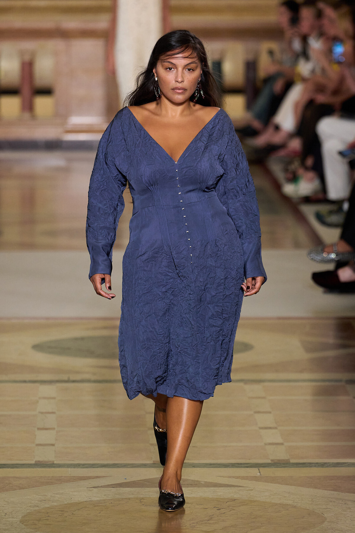 Model walks a runway in a blue crinkled midi dress with a deep V-neck and front buttons, sleeves full and cuffs.
